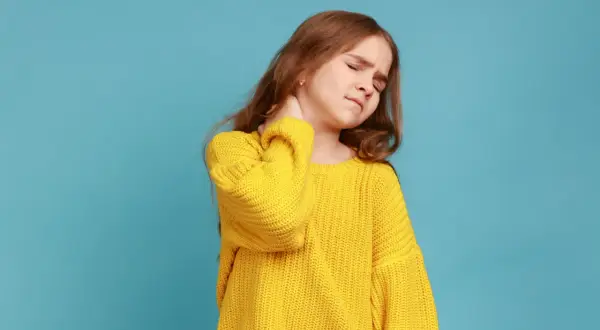 Ein Mädchen mit einem gelben Pullover hält sich den Nacken. Sie hat einen steifen Nacken und sieht gequält aus.