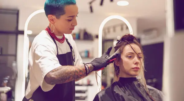 Friseurin mit blauen Haaren blondiert die Haare einer Kundin. Die Kundin wartet geduldig auf das Ergebnis, während die Friseurin sorgfältig arbeitet.
