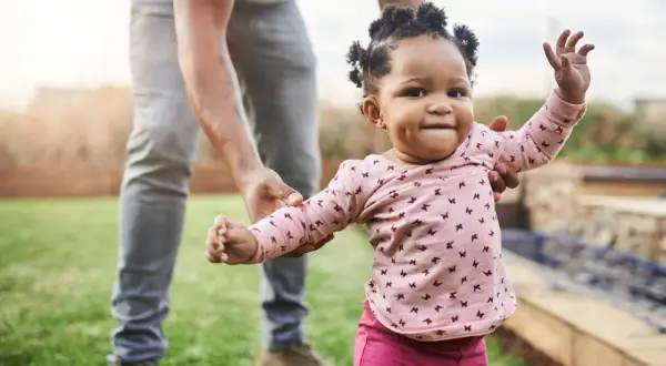 Ein Baby lernt laufen! Ein kleines Mädchen in einem rosa Shirt mit Schmetterlingen macht seine ersten Schritte im Garten, gehalten von einer erwachsenen Hand.