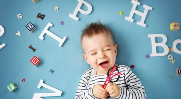 Lachendes Baby mit Brille inmitten von Buchstaben. Bald beginnt die spannende Reise, ab wann sprechen Kinder?