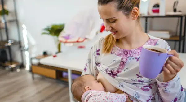 Mutter stillt Baby, trinkt Kaffee. Mögliche baby koffein symptome beachten.