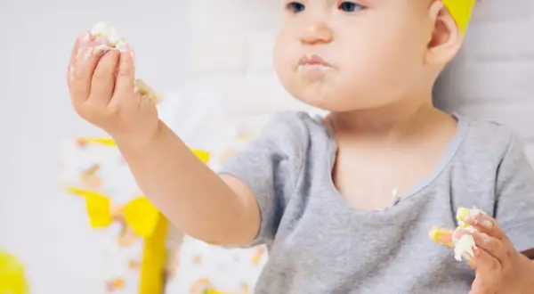 Kleines Kind mit gelbem Haarband isst mit den Händen, das Gesicht ist voller Polenta.