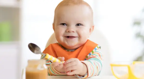 Glückliches Baby mit Lätzchen und Löffel, bereit für die Beikost. Zeichen der Freude beim Essen.