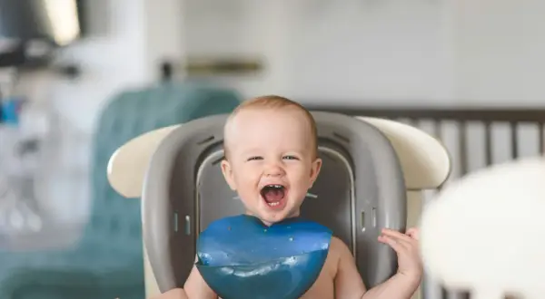 Ein glückliches Baby mit einem blauen Lätzchen sitzt im Hochstuhl und freut sich über seinen ersten Brei.