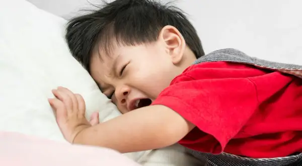 Ein weinendes Baby haut sich auf den Kopf, liegt auf einem Kissen und trägt ein rotes Oberteil.