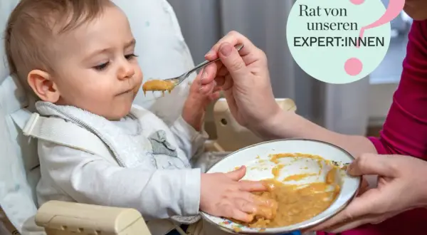 Ein Baby sitzt im Hochstuhl und bekommt Brei gefüttert. Die Hände des Babys sind voller Essen. Tipps für Baby essen von Expert:innen.