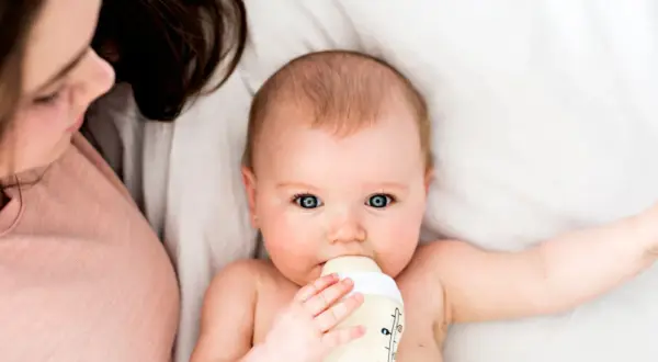 Ein Baby trinkt aus einer Flasche, während eine Person daneben liegt. Die Frage, wie lange braucht Essen bis es beim Baby ankommt, beschäftigt viele Eltern.