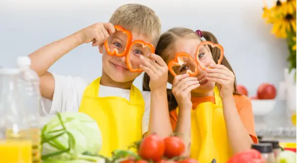 Zwei Kinder mit Paprikaringen vor den Augen, die Spaß beim Kochen haben. Ein wichtiger Aspekt für gesunde Entwicklung ist der richtige kalorienbedarf kind 10 jahre.