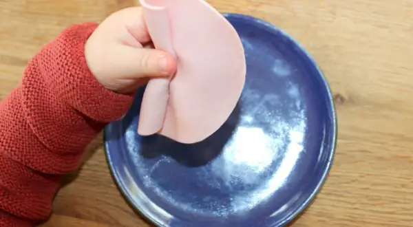 Kleines Baby mit rotem Pullover hält Wurstscheibe über blauen Teller. Ab wann darf Baby Wurst essen?