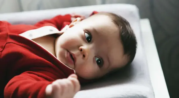 Ein süßes Baby in rotem Strampler liegt auf einer Wickelauflage. Es schaut neugierig in die Kamera, als ob es gerade vom Wickeltisch gefallen wäre und sich wundert, wie es hierher kam.