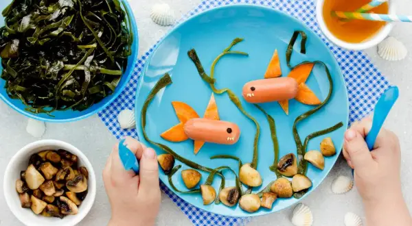 Kreativer Fischbrei für Babys: Würstchen-Fische schwimmen im Meer aus Algen und Pilzen auf einem blauen Teller.