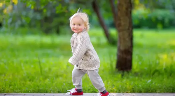 Ein fröhliches, blondes Kind, circa 2 Jahre alt, läuft auf einem Weg im Park. Es trägt einen Strickcardigan und rote Turnschuhe.