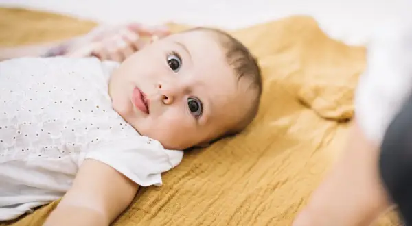 Ein neugieriges Baby, etwa 3 Monate alt, liegt auf einer senfgelben Decke und blickt mit großen Augen in die Kamera.