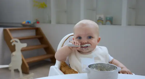 Kleines Baby mit blauen Augen isst begeistert Baby Naturjoghurt. Löffel und Lätzchen sind voller leckerer Reste.