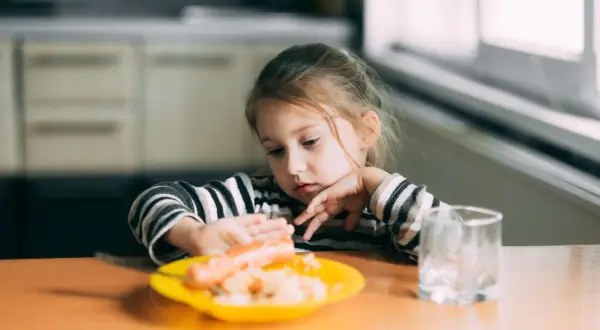 Ein kleines Mädchen mit nachdenklichem Blick betrachtet eine Wurst auf ihrem Teller. Typisches Essverhalten Kinder.