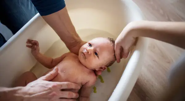 Eltern helfen ihrem Baby beim Duschen in einer Babybadewanne. Sanfte Hände halten das Kleine, während es im Wasser planscht.