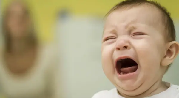 Ein Baby kreischt schrill, die Augen geschlossen, der Mund weit geöffnet. Im Hintergrund eine unscharfe Person.