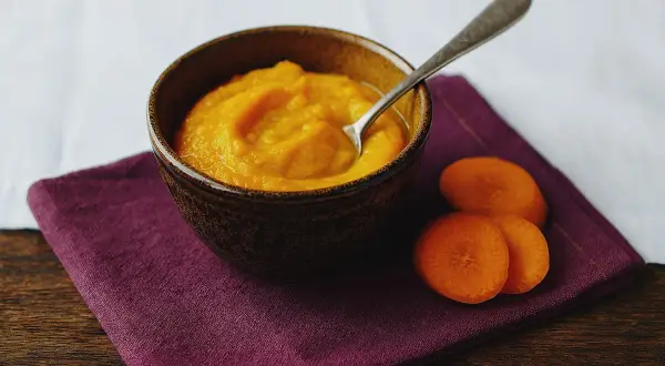 Ein Schälchen mit orangefarbenem Babybrei, daneben Karottenscheiben. Perfekt für den ersten Fisch Babybrei.