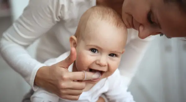Ein Baby mit einem Daumen, der sanft die Zähne berührt, um die Zahnen Symptome zu lindern. Die Mutter küsst das Kind liebevoll.