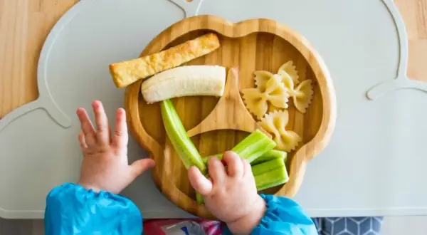 Baby obst ab wann? Ein Kleinkind greift nach Banane, Gurke und Nudeln auf einem Bambusteller.
