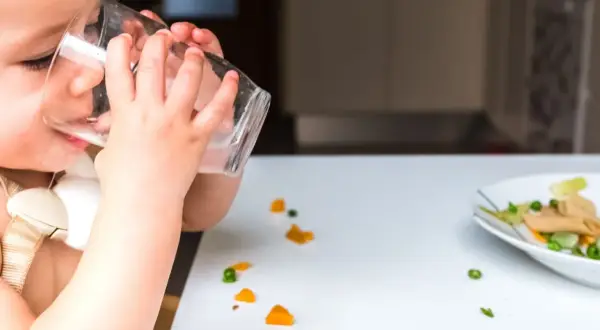 Kleines Kind trinkt aus Glas, daneben ein Teller mit Brei und verstreuten Gemüsestückchen. Wie viel Wasser bei Beikost?