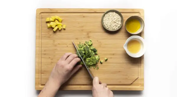 Hände schneiden Brokkoli für Babybrei. Kartoffelwürfel, Haferflocken und Öl sind bereit.
