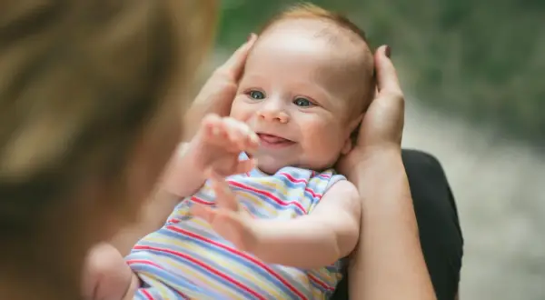Ein lächelndes neugeborenes Baby mit blauen Augen wird von Händen gehalten. Es trägt ein gestreiftes Oberteil und streckt seine Arme aus.