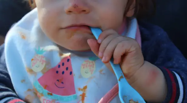 Ein Baby mit Lätzchen isst mit einem blauen Löffel. Das Lätzchen zeigt Früchte und das Wort "Fruchtchen". Das ist ein Beispiel für Beikost.