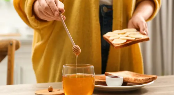 Goldener Honig tropft von einem Holzstäbchen auf ein Glas. Eine Person bereitet Toast mit Bananen und Honig zu, ein süßer Genuss beim Stillen.