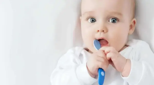 Ein Baby mit großen blauen Augen hält einen blauen Löffel und steckt ihn in den Mund. Es ist ein süßer sprung 5 baby.