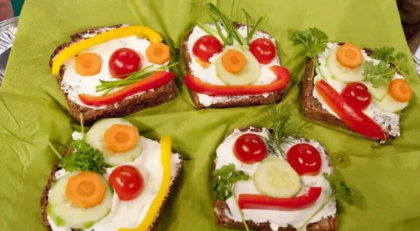Lustige Gesichter auf belegten Broten: Fingerfood, das Kinder lieben. Bunte Gemüsesorten wie Paprika, Gurke und Karotte schmücken die Brotscheiben.