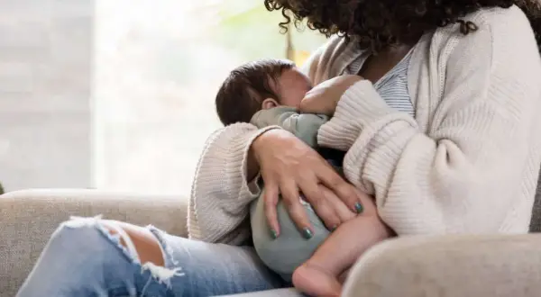 Mutter stillt unruhiges Baby, während sie auf einem Sofa sitzt. Ein Kaffee steht vielleicht in Reichweite.