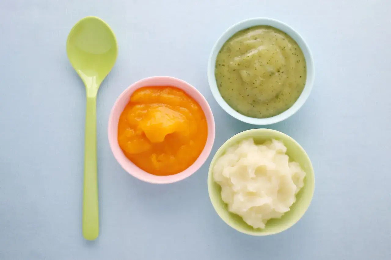 Drei Schälchen mit Babynahrung: orangefarben, grün und weiß. Welcher Joghurt Baby ist wohl am leckersten? Ein grüner Löffel liegt daneben.