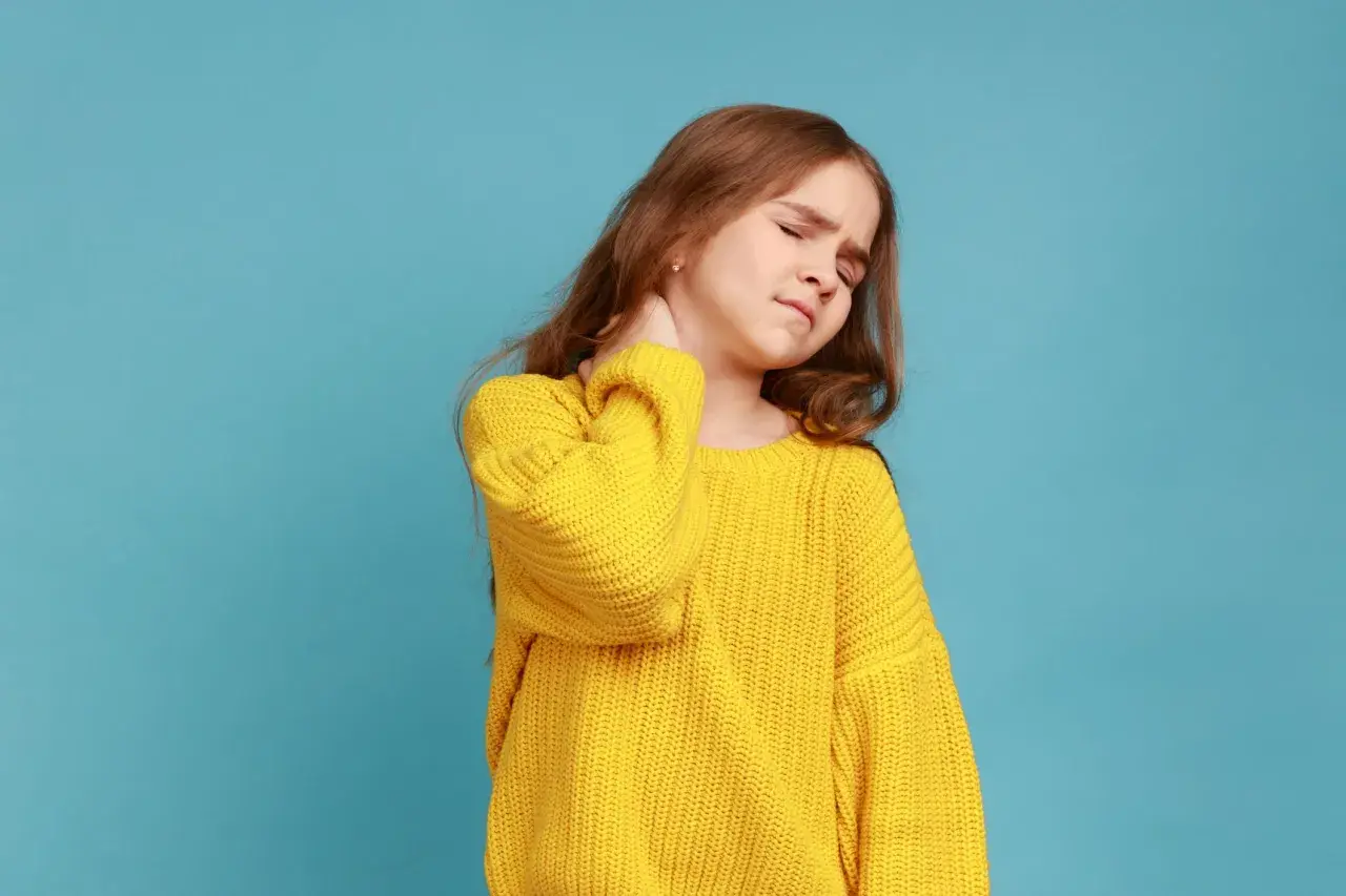 Ein Mädchen mit einem gelben Pullover hält sich den Nacken. Sie hat einen steifen Nacken und sieht gequält aus.