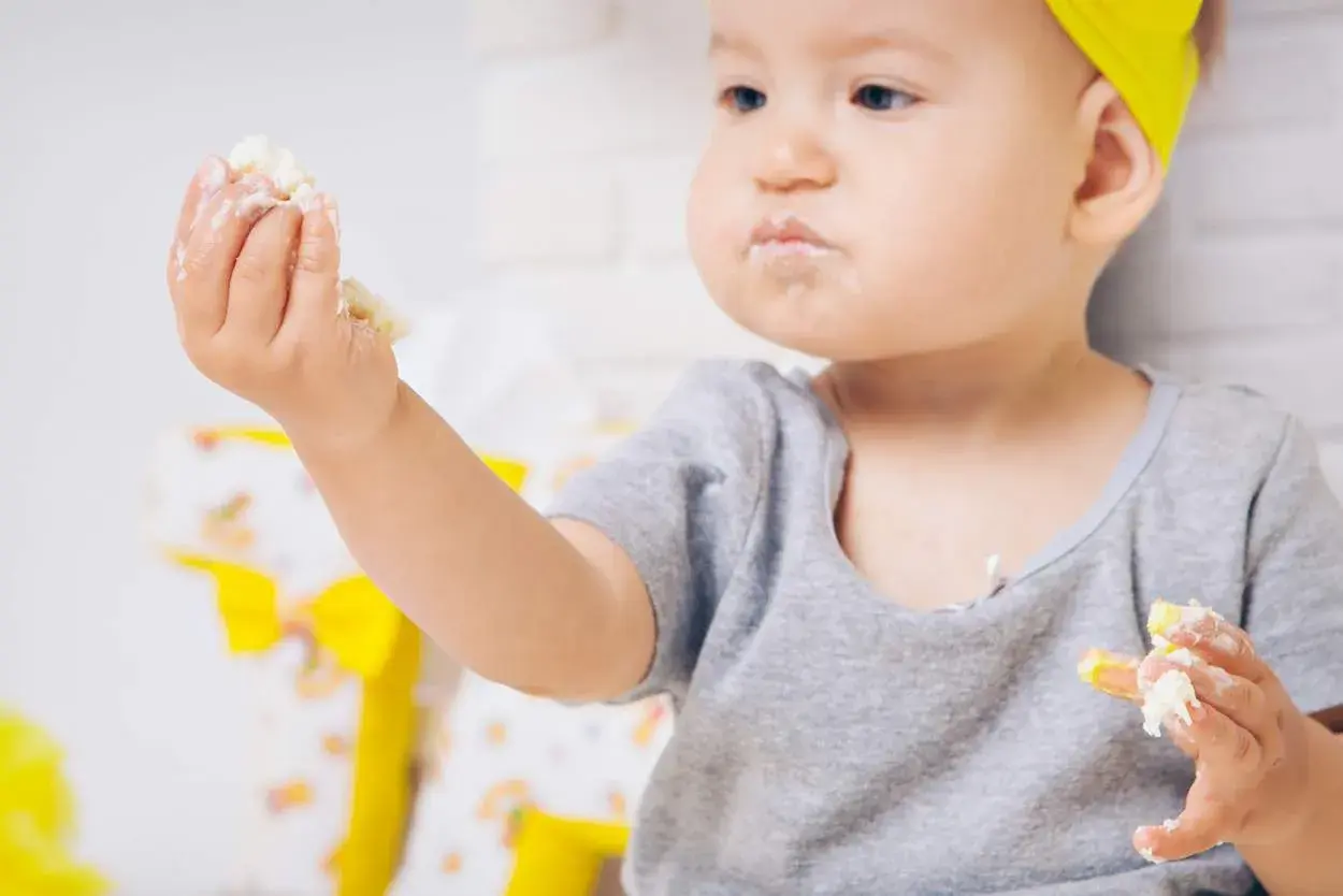 Kleines Kind mit gelbem Haarband isst mit den Händen, das Gesicht ist voller Polenta.