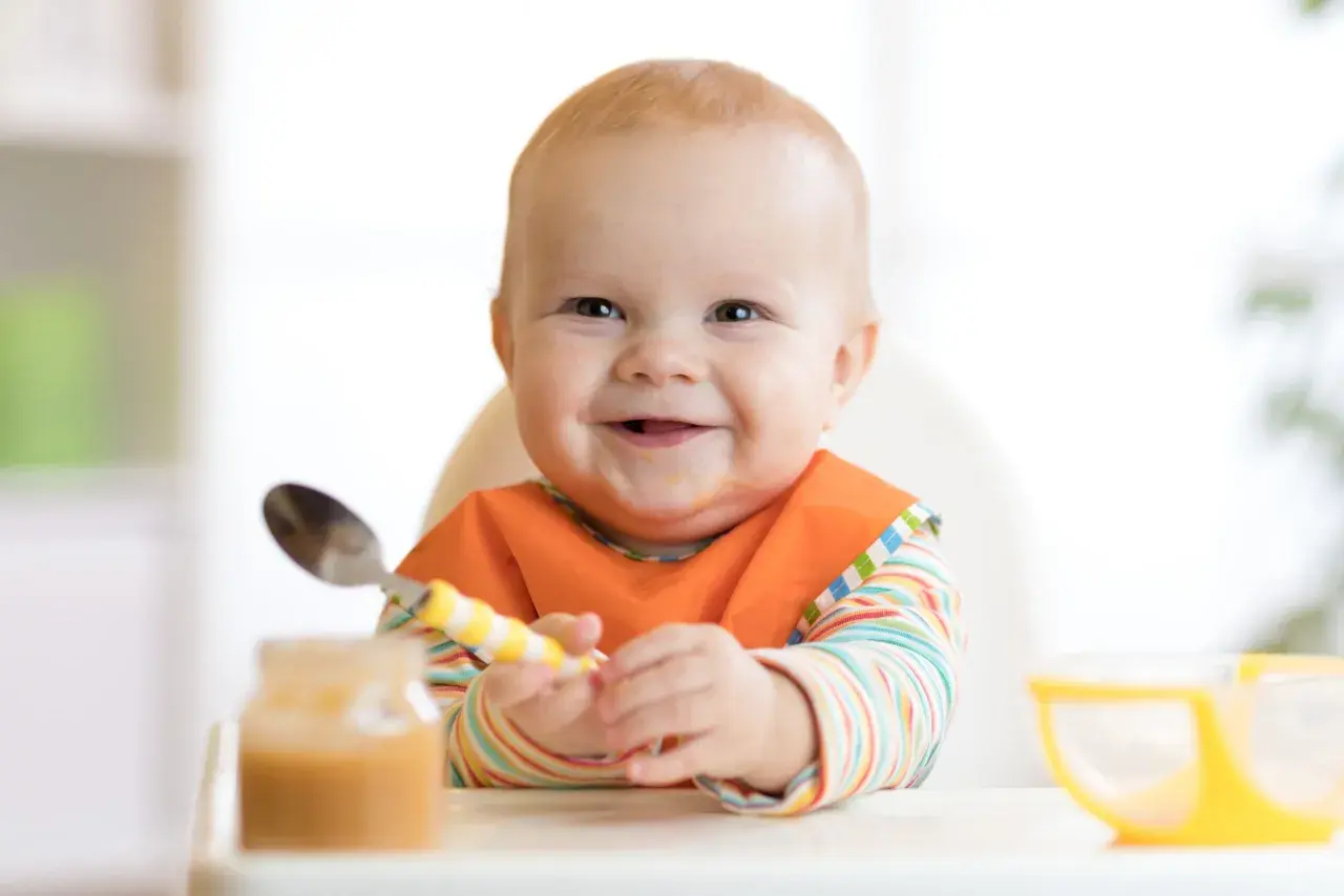 Glückliches Baby mit Lätzchen und Löffel, bereit für die Beikost. Zeichen der Freude beim Essen.