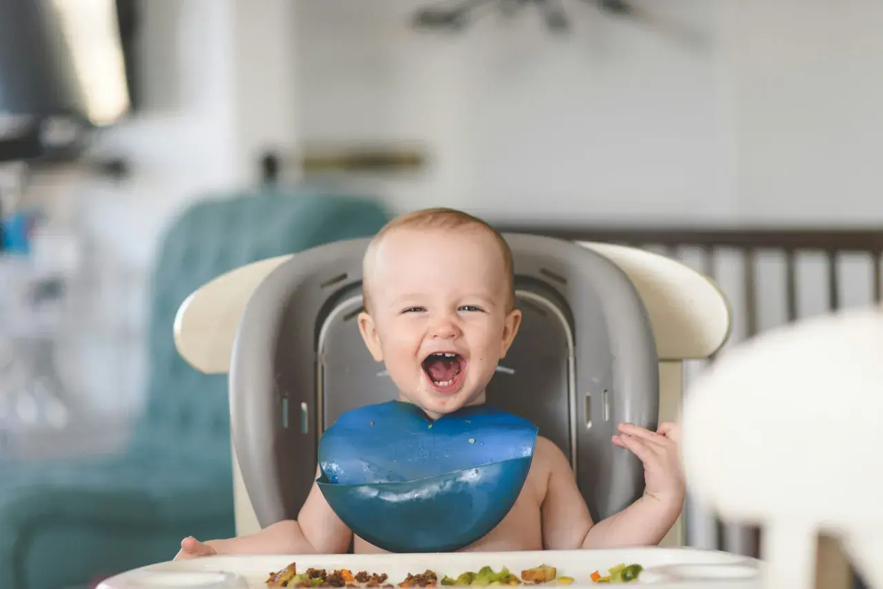 Ein glückliches Baby mit einem blauen Lätzchen sitzt im Hochstuhl und freut sich über seinen ersten Brei.