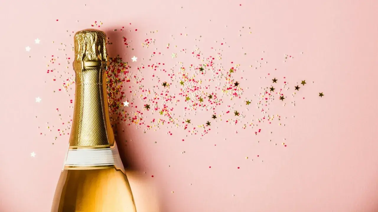 Goldene Sektflasche mit bunten Streuseln und Sternen auf rosa Hintergrund. Ein Glas Sekt stillen für den besonderen Moment.
