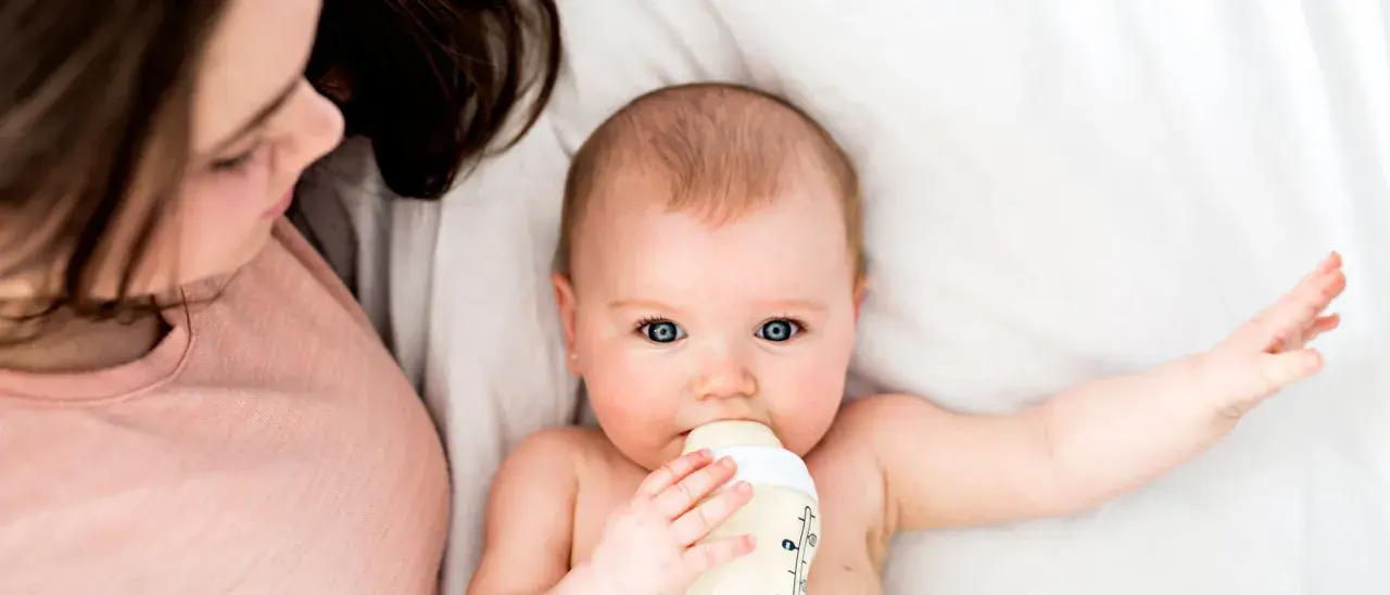Ein Baby trinkt aus einer Flasche, während eine Person daneben liegt. Die Frage, wie lange braucht Essen bis es beim Baby ankommt, beschäftigt viele Eltern.