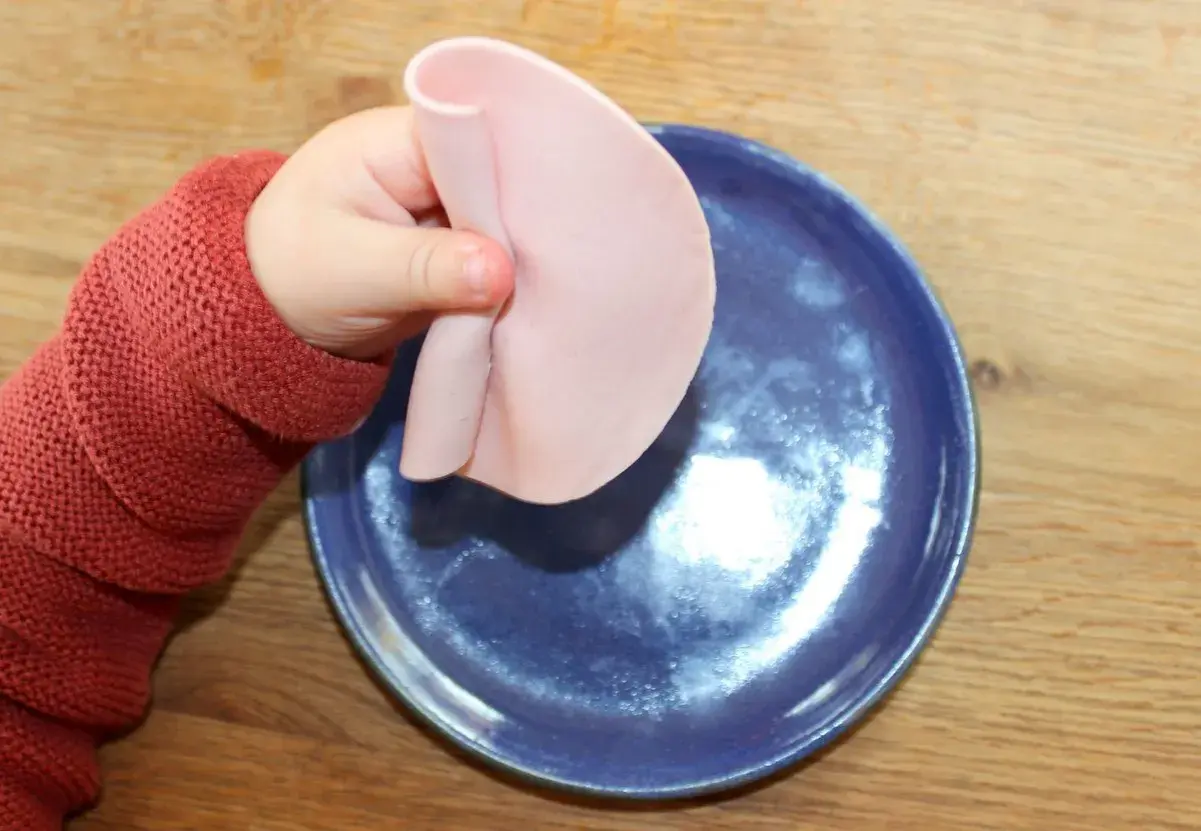 Kleines Baby mit rotem Pullover hält Wurstscheibe über blauen Teller. Ab wann darf Baby Wurst essen?