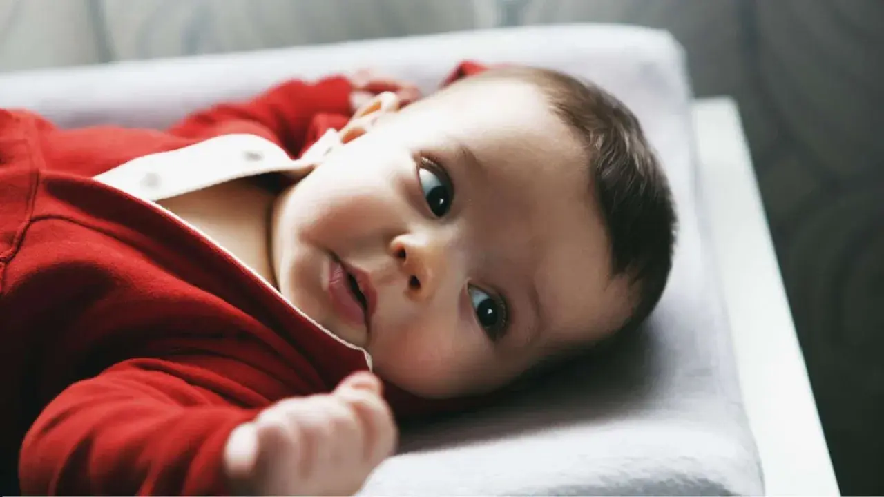 Ein süßes Baby in rotem Strampler liegt auf einer Wickelauflage. Es schaut neugierig in die Kamera, als ob es gerade vom Wickeltisch gefallen wäre und sich wundert, wie es hierher kam.