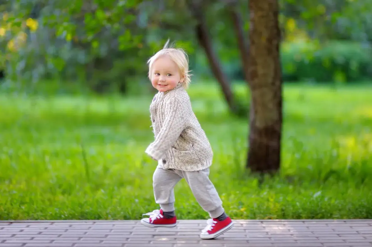 Ein fröhliches, blondes Kind, circa 2 Jahre alt, läuft auf einem Weg im Park. Es trägt einen Strickcardigan und rote Turnschuhe.