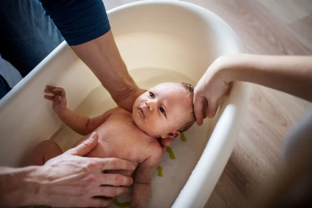 Eltern helfen ihrem Baby beim Duschen in einer Babybadewanne. Sanfte Hände halten das Kleine, während es im Wasser planscht.
