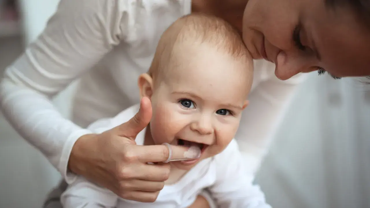 Ein Baby mit einem Daumen, der sanft die Zähne berührt, um die Zahnen Symptome zu lindern. Die Mutter küsst das Kind liebevoll.