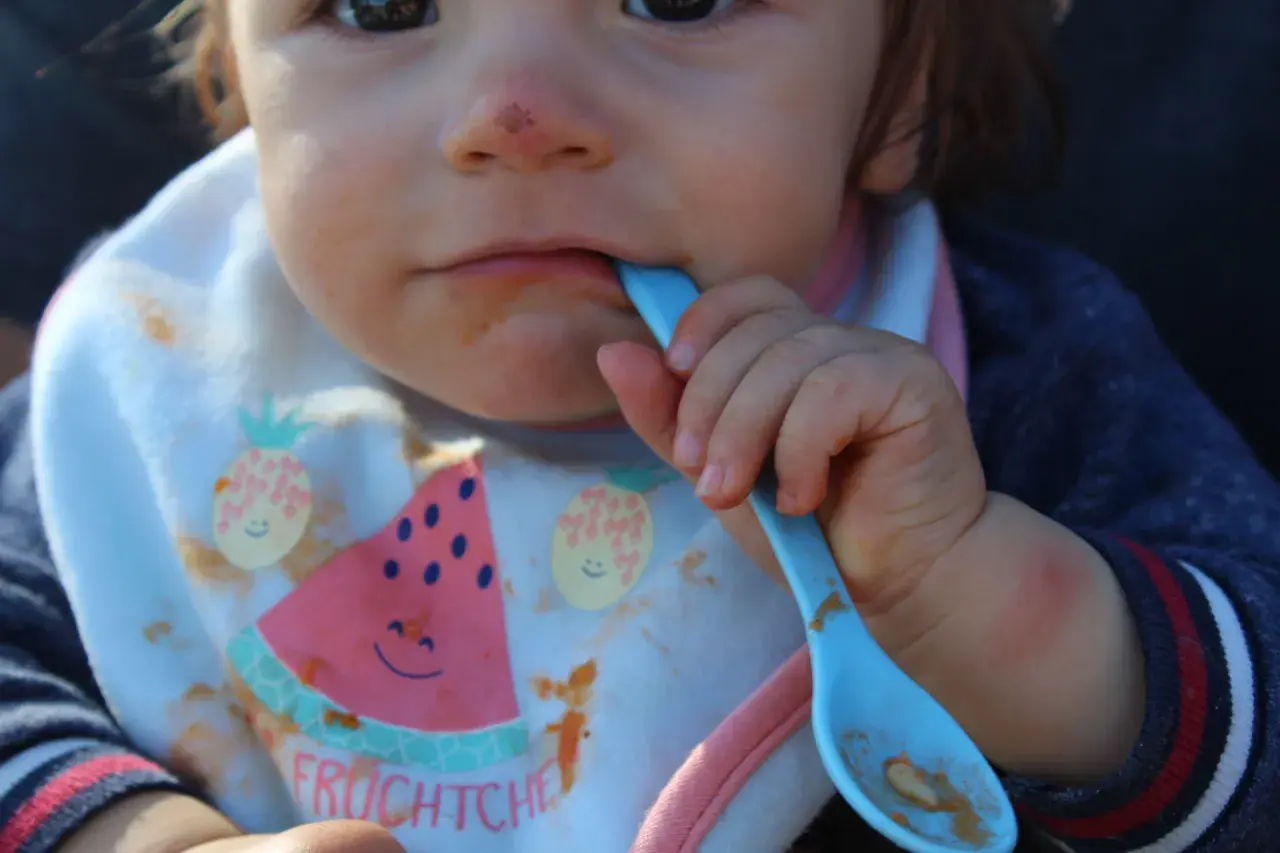 Ein Baby mit Lätzchen isst mit einem blauen Löffel. Das Lätzchen zeigt Früchte und das Wort "Fruchtchen". Das ist ein Beispiel für Beikost.