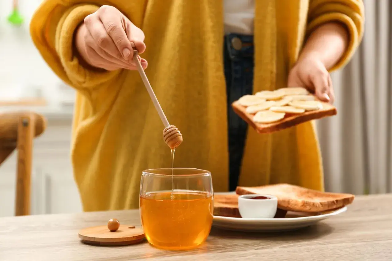 Goldener Honig tropft von einem Holzstäbchen auf ein Glas. Eine Person bereitet Toast mit Bananen und Honig zu, ein süßer Genuss beim Stillen.