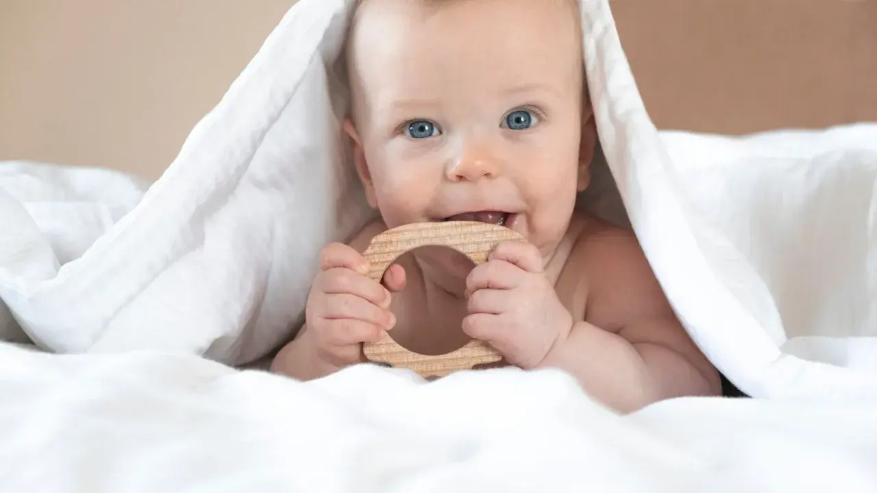 Blauäugiges Baby unter weißer Decke, kaut auf Holzring. Ab wann sabbern Babys? Oft schon vor dem Zahnen.