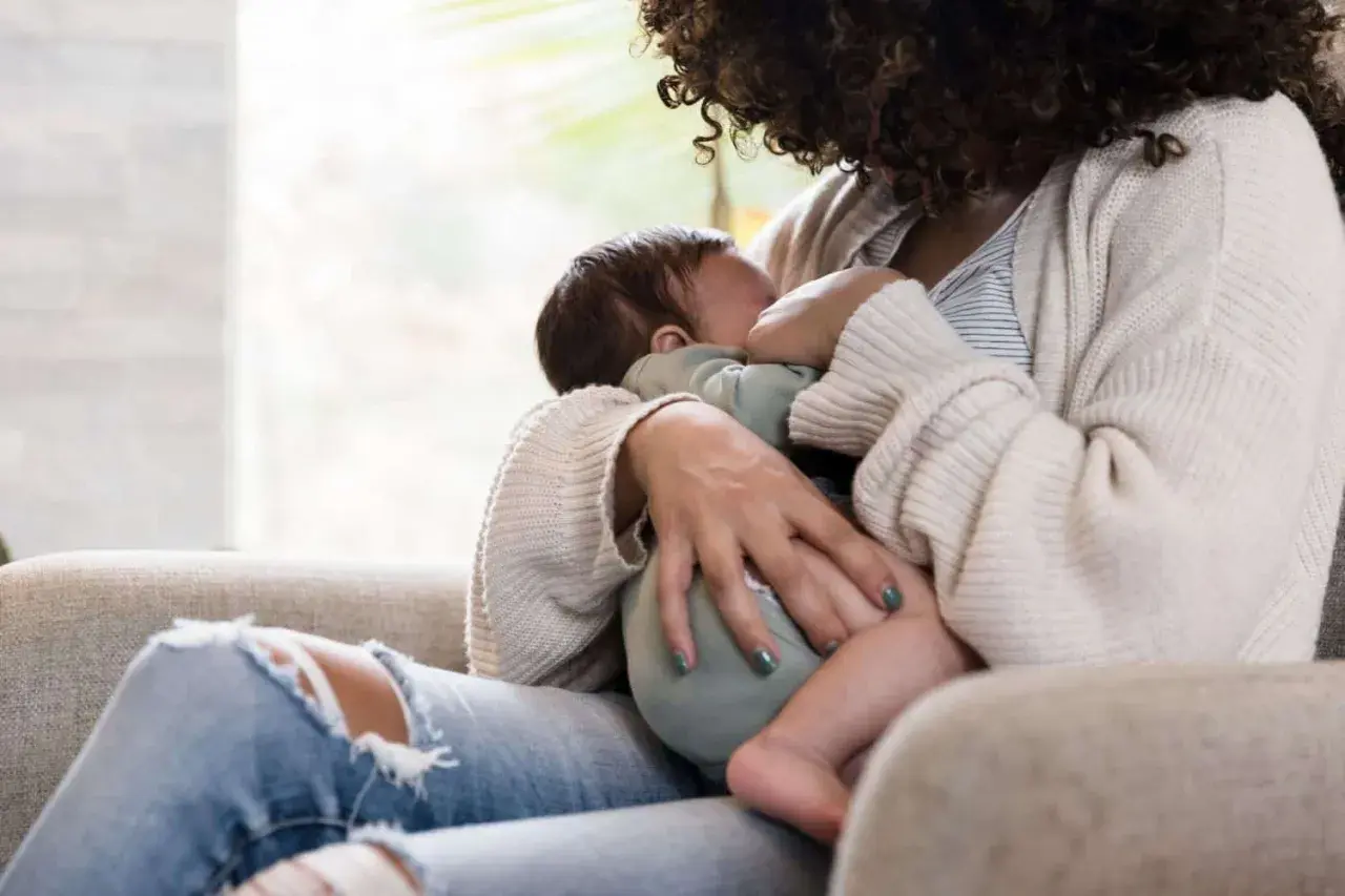 Mutter stillt unruhiges Baby, während sie auf einem Sofa sitzt. Ein Kaffee steht vielleicht in Reichweite.