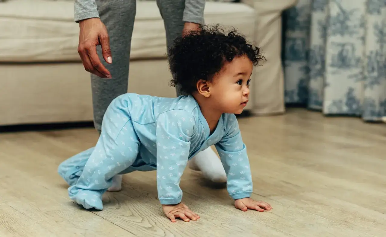 Kleines Baby in blauem Strampler krabbelt auf dem Boden, wedelt mit Armen und Beinen. Eine Hand hält es sanft.