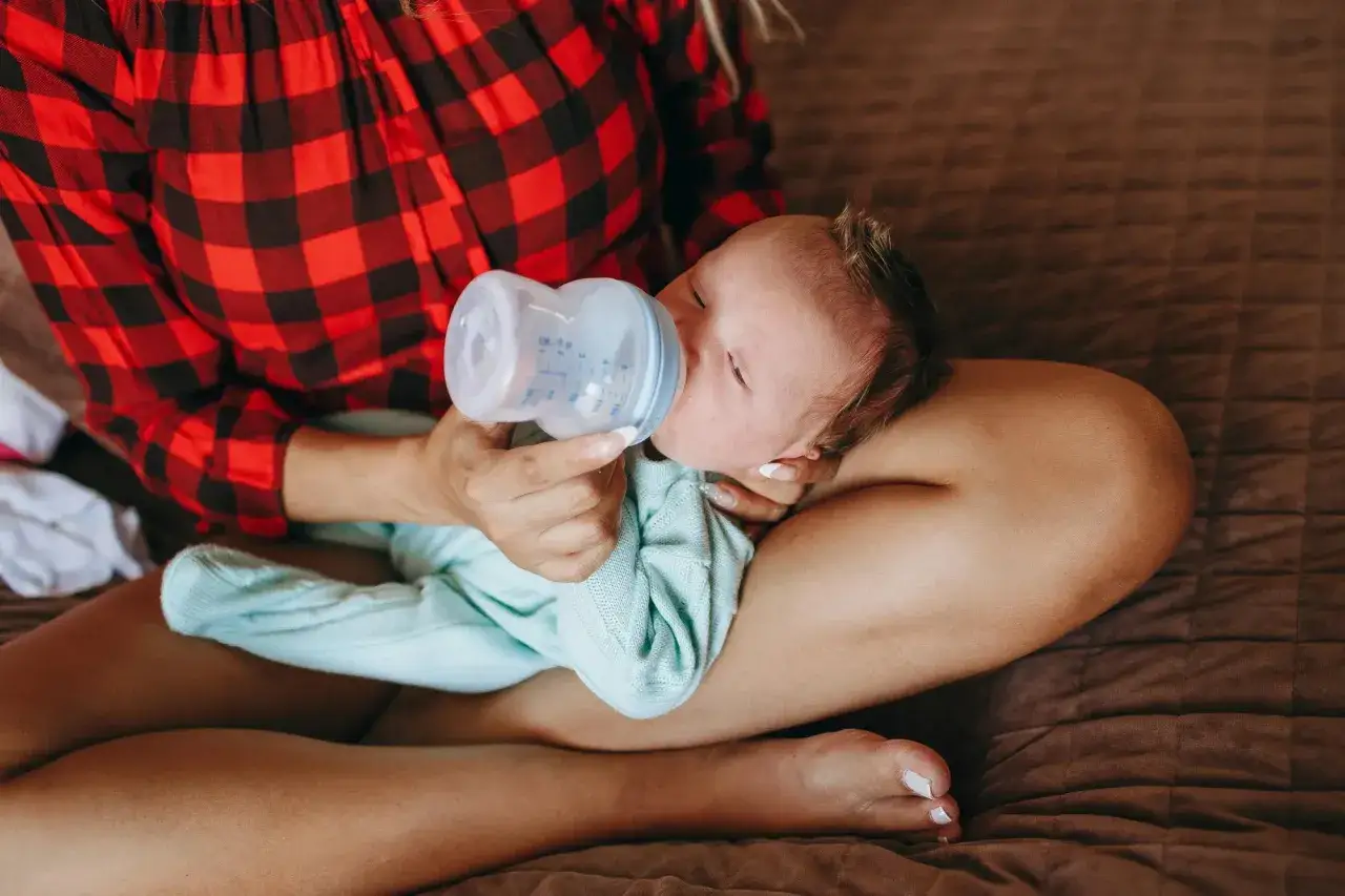 Ein Baby wird mit einer Flasche gef&uuml;ttert, w&auml;hrend es auf dem Scho&szlig; einer Person liegt. Die Person tr&auml;gt ein rot-schwarzes kariertes Hemd.